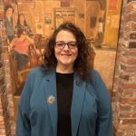 Joanne wears a blue blazer. She has brown hair and glasses. She smiles at the camera against a brick background.