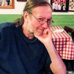 Joel Deuel is pictured with glasses and brown hair pulled back in a ponytail. He's wearing a black shirt and is sitting in a restaurant.