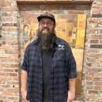 Mateo smiles at the camera against a brick background. He wears a blue and black flannel and a baseball hat.