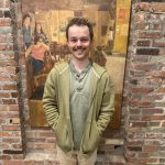 Reese is pictured in front of a brick wall. He smiles at the camera. He is in a light green jacket and has brown hair.